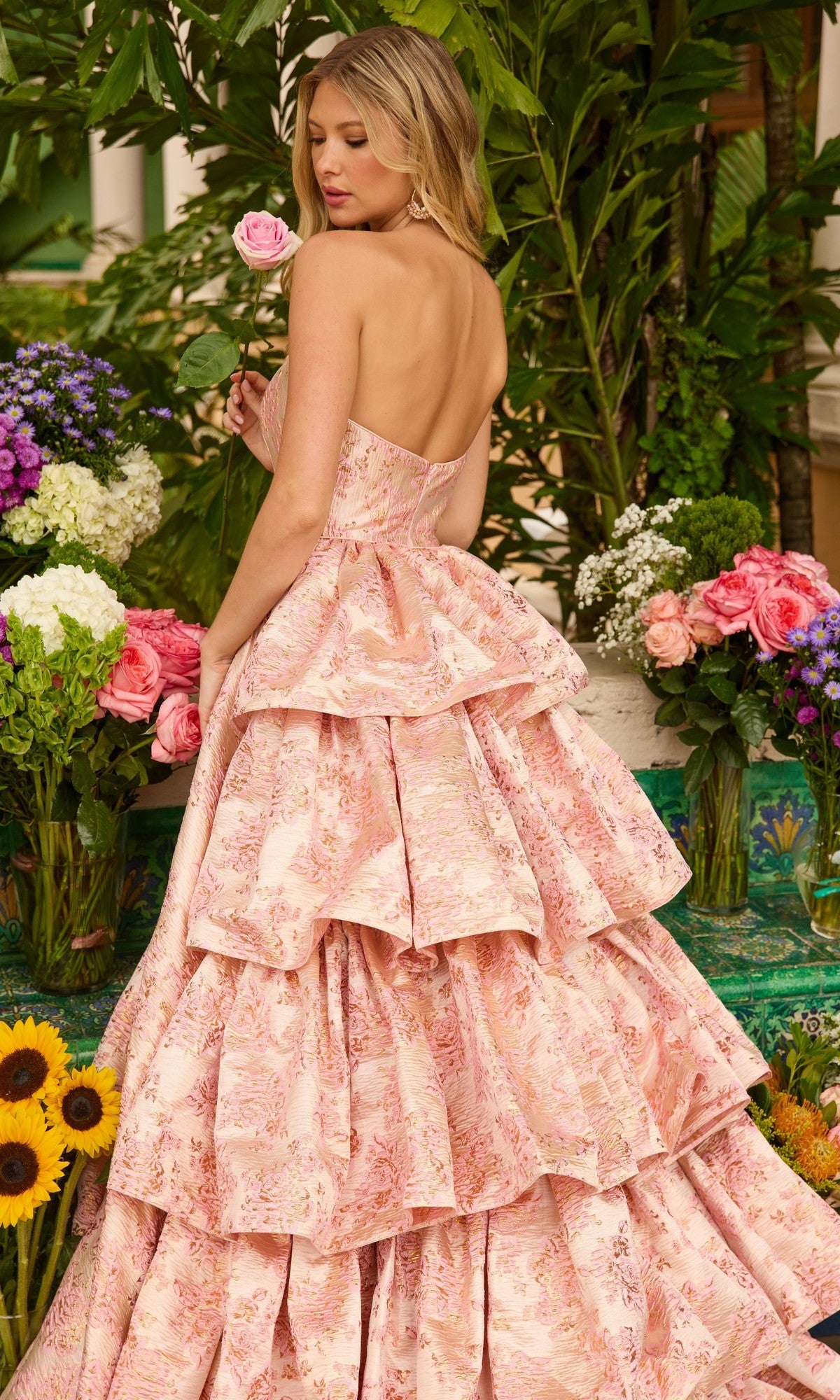 A woman in the Ava Presley 26412 Ruffled Strapless Prom Gown stands among vibrant flowers, holding a pink rose and glancing over her shoulder in her floral print evening gown.